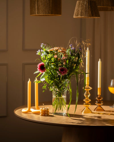 Ein Blumenstrauß in einer Glasvase "So kunterbunt wie Du" steht auf einem runden Holztisch, umgeben von dekorativen Kerzen und Kerzenständern, die in warmes Licht getaucht sind.