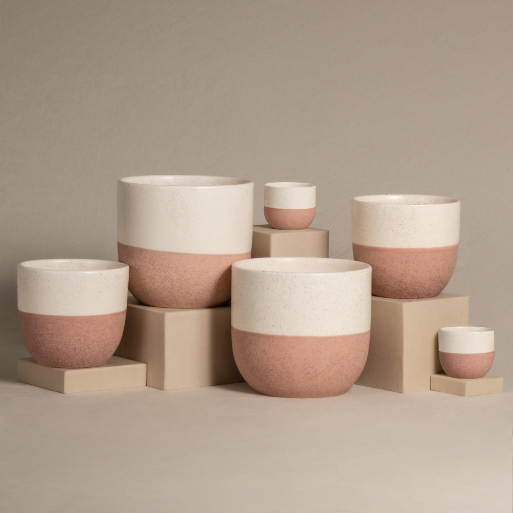 Das handgefertigte Keramik-Topfset „Variado“ zeichnet sich durch ein minimalistisches Design mit einer weißen oberen Hälfte und einer terrakottafarbenen unteren Hälfte aus. Diese wunderschön gefertigten Innentöpfe werden auf beigen Plattformen unterschiedlicher Höhe vor einem neutralen Hintergrund präsentiert.