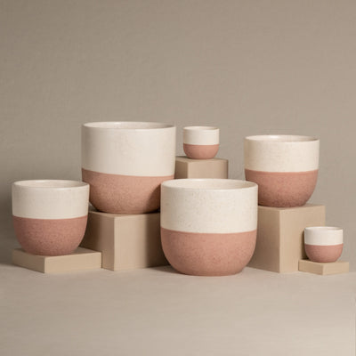 Das handgefertigte Keramik-Topfset „Variado“ zeichnet sich durch ein minimalistisches Design mit einer weißen oberen Hälfte und einer terrakottafarbenen unteren Hälfte aus. Diese wunderschön gefertigten Innentöpfe werden auf beigen Plattformen unterschiedlicher Höhe vor einem neutralen Hintergrund präsentiert.