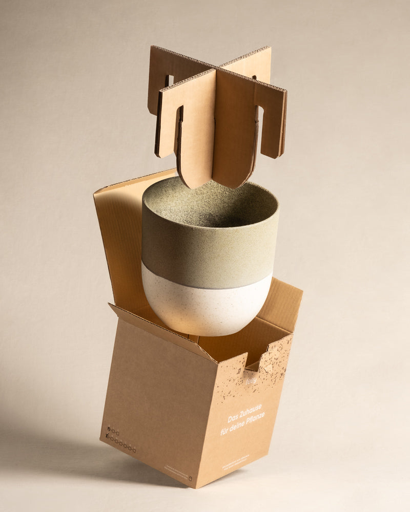 Ein Keramik-Topfset „Variado“ (22, 16, 14) mit zweifarbigem Design (grünlich-grau und weiß) ist teilweise aufgehängt und von einer braunen Kartonverpackung umgeben, die es schützen soll. Die Schachtel ist leicht geöffnet und zeigt so ihren einzigartigen und sicheren Verpackungsmechanismus.
