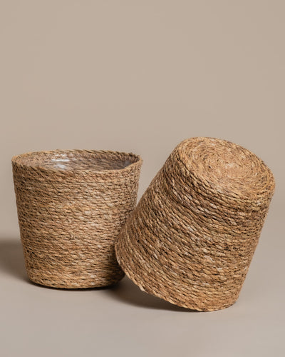 Zwei geflochtene "Pflanzenkörbe aus Seegras (Kesat | 18 cm ⌀)" ruhen auf weißem Grund, jeder ein handgeflochtenes Einzelstück mit eigener Textur und eigenem Charme.