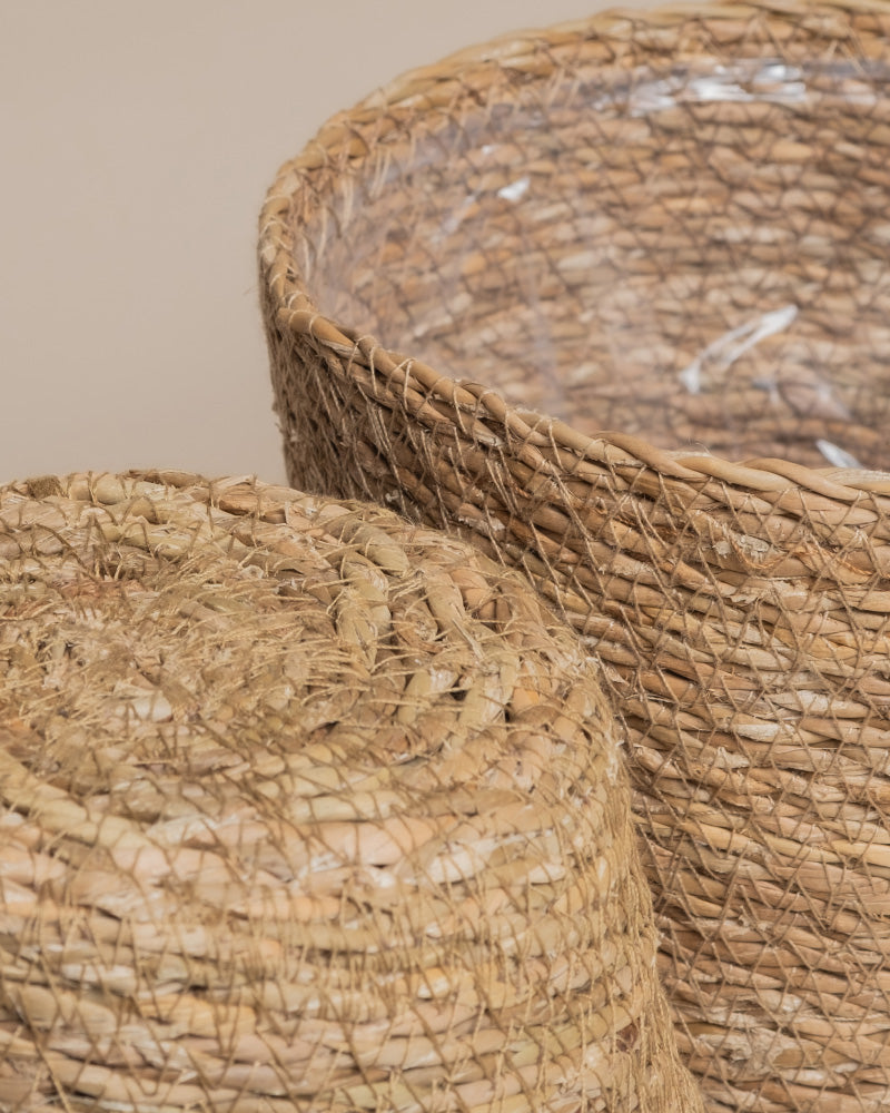 Nahaufnahme von zwei geflochtenen, runden Pflanzenkorb-Sets aus Seegras (Kesat | 2× 16) mit rauer Textur. Der größere Korb, aufrecht und mit durchsichtigem Kunststoff ausgekleidet, und der kleinere, umgedreht, stehen vor einem neutralen beigen Hintergrund.