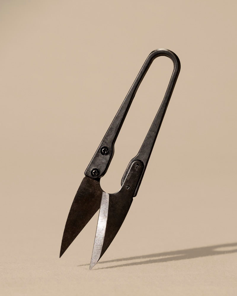 Eine Fadenschere der Marke Kleine Pflanzenschere aus dunklem Kohlenstoffstahl steht aufrecht auf einer neutralen beigen Oberfläche mit leicht geöffneten Klingen und einem einfachen, zweckmäßigen Design und wirft einen Schatten nach unten.