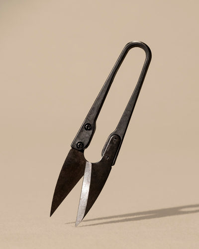 Eine Fadenschere der Marke Kleine Pflanzenschere aus dunklem Kohlenstoffstahl steht aufrecht auf einer neutralen beigen Oberfläche mit leicht geöffneten Klingen und einem einfachen, zweckmäßigen Design und wirft einen Schatten nach unten.