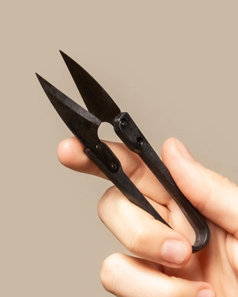 Eine Hand hält eine kleine Pflanzenschere mit scharfen, spitzen Klingen vor einem schlichten beigen Hintergrund.