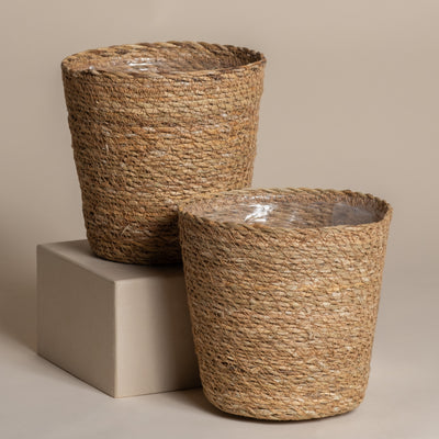 Zwei naturfarbene Pflanzenkorb-Sets aus Seegras (Kesat | 2 × 18) mit Kunststoffeinlagen - eines auf einem hellbeigen Block, eines auf dem Boden - werden vor einem neutralen Hintergrund elegant in Szene gesetzt.