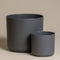 Zwei moderne, minimalistisch anmutende dunkelgraue Blumentöpfe des Kunststoff-Topfsets „Lumi“ in unterschiedlichen Größen stehen nebeneinander auf einer beigen Oberfläche. Der größere Topf (25 cm) steht links, der kleinere (14 cm) rechts. Der Hintergrund ist in einem neutralen Beige gehalten, perfekt für jeden Innenbereich.