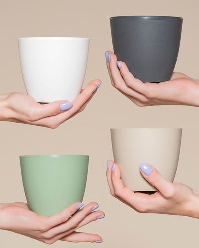 Vier Hände mit hellviolettem Nagellack halten jeweils einen andersfarbigen Kunststoff-Topf (Kiki | 11 cm Ø) in Weiß, Dunkelgrau, Hellgrün und Beige - die perfekte Zuhause Deko, um Ihr Pflänzchen vor einem neutralen beigen Hintergrund in Szene zu setzen.