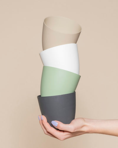 Eine Hand mit hellviolettem Nagellack hält vier gestapelte Kunststoff-Töpfe (Kiki | 11 cm Ø) in gedämpftem Grau, Grün, Weiß und Beige vor einem beigen Hintergrund - perfekt für eine stilvolle Wohndekoration.