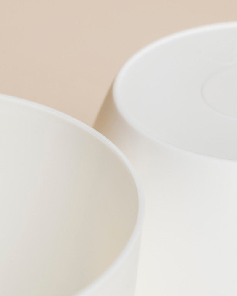Nahaufnahme von zwei weißen, runden Kunststoff-Töpfen (Kiki | 11 cm Ø) auf einem neutralen beigen Hintergrund, wobei ihre partiellen Ränder und glatten Oberflächen unter sanfter Beleuchtung hervorgehoben werden.