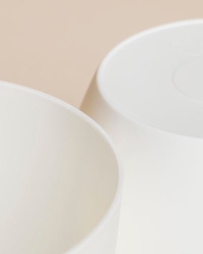 Nahaufnahme von zwei weißen, runden Kunststoff-Töpfen (Kiki | 11 cm Ø) auf einem neutralen beigen Hintergrund, wobei ihre partiellen Ränder und glatten Oberflächen unter sanfter Beleuchtung hervorgehoben werden.