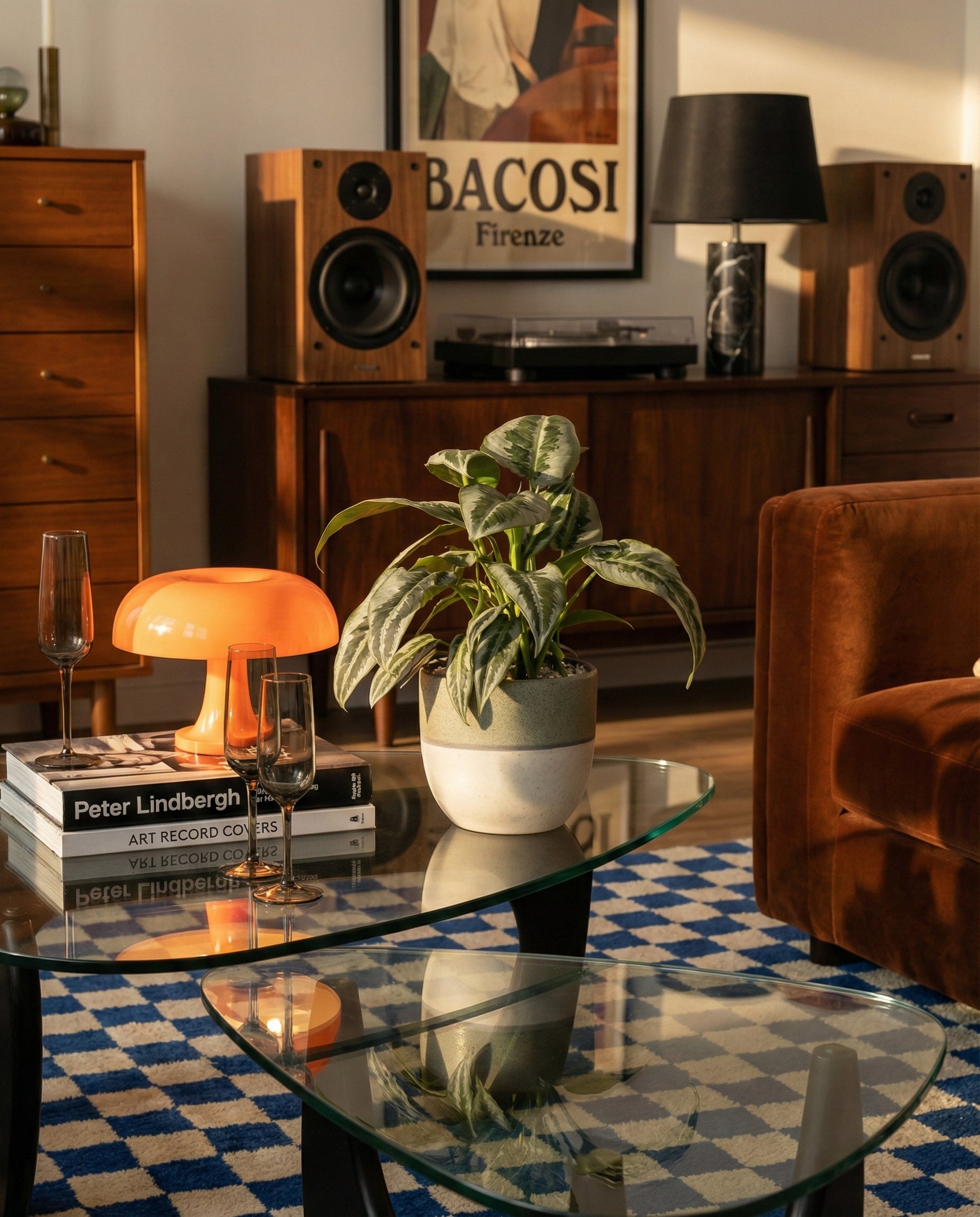 Sonnendurchflutetes Wohnzimmer mit einem gläsernen Couchtisch, einer Schismatoglottis 'Silver Indukan' - feey-Topfpflanze, einer orangefarbenen Pilzlampe, gestapelten Büchern und Sektgläsern. Im Hintergrund eine hölzerne Stereokonsole mit Lautsprechern und gerahmter Kunst an der Wand.