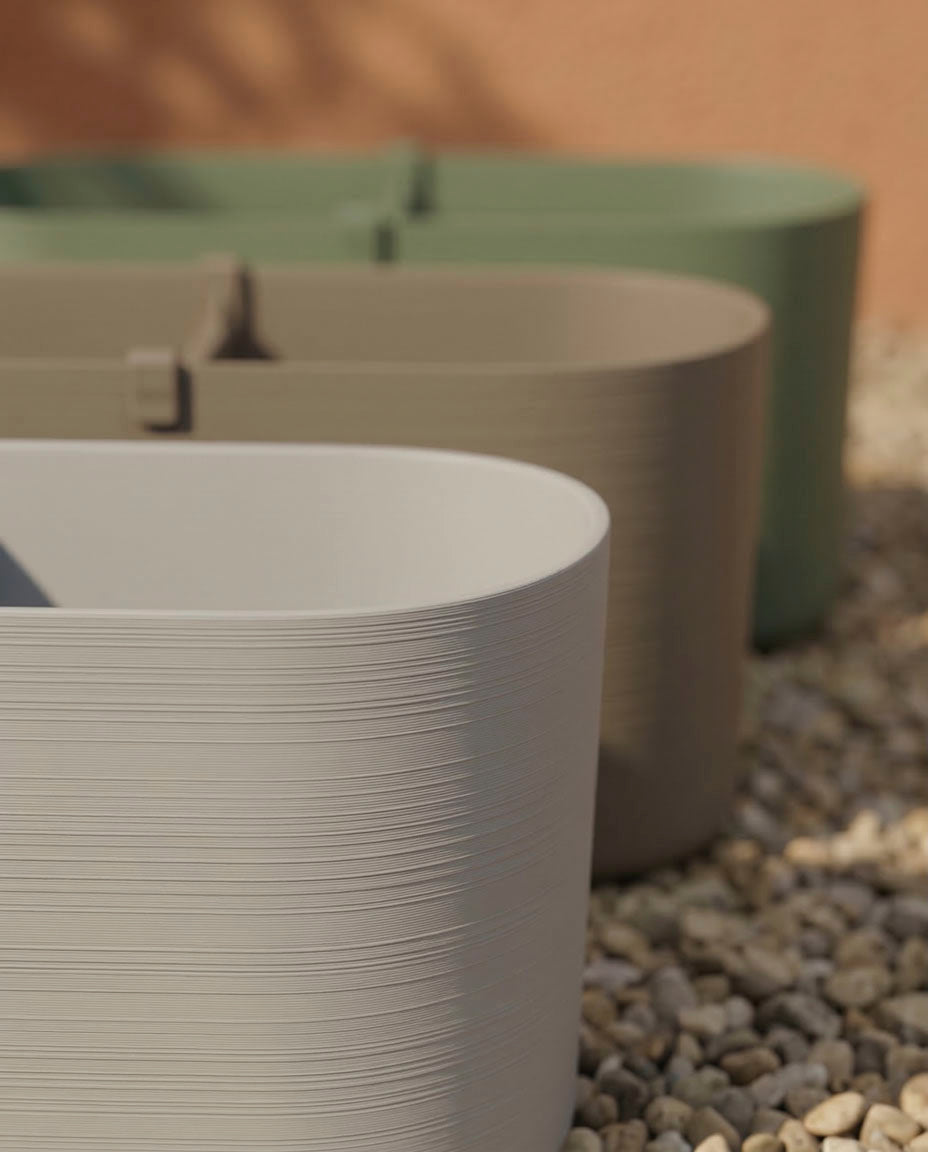 Drei große, ovale Pflanzkästen (Paros | 60 cm) in den Farben Beige, Taupe und Grün sind auf Kieselsteinen angeordnet - ideal für Balkonpflanzen mit eingebauter Drainage, um die Pflanzen gesund zu halten, während das Sonnenlicht sanfte Hintergrundschatten erzeugt.