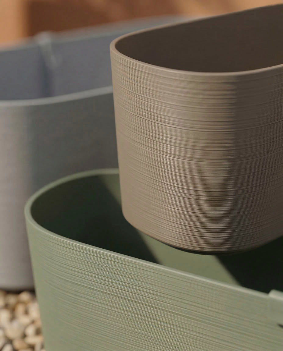 Drei ovale Pflanzkästen (Paros | 60 cm) in gedämpftem Grau, Taupe und Salbei sind aufeinander gestapelt - perfekt für Balkonpflanzen. Das Sonnenlicht hebt ihre strukturierten Rippen hervor, und ein Boden aus weißen Kieselsteinen deutet auf eine hervorragende Drainage hin.
