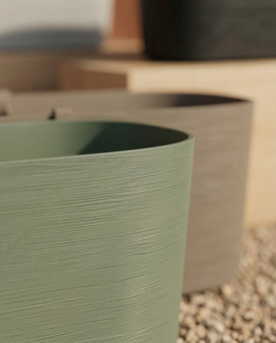 Nahaufnahme eines strukturierten, ovalen Pflanzkastens (Paros | 60 cm) mit Drainage in gedämpftem Grün und Beige, der auf Kieselsteinen steht. Im Hintergrund sind stilvolle Pflanzgefäße in sanften, neutralen Tönen zu sehen.