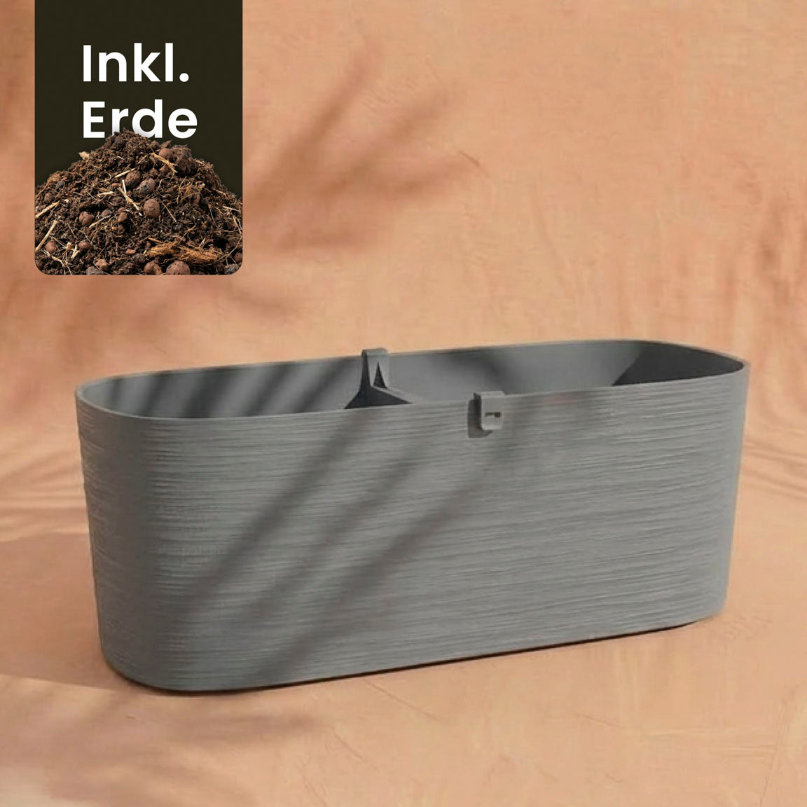 Der Pflanzkasten (Paros | 60 cm) ist ein großes, rechteckiges graues Pflanzgefäß auf pfirsichfarbener Oberfläche. Ein Einsatz zeigt Erde mit 