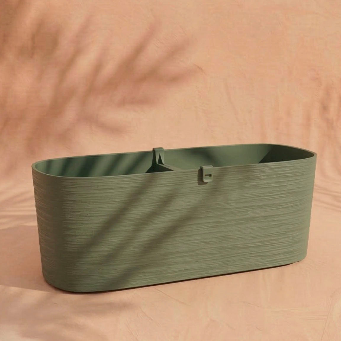 Der Pflanzkasten (Paros | 60 cm) ist ein grünes, ovales Pflanzgefäß mit strukturierter Oberfläche und eingebauter Drainage, das auf einem zart pfirsichfarbenen Hintergrund mit Blattschatten abgebildet ist.