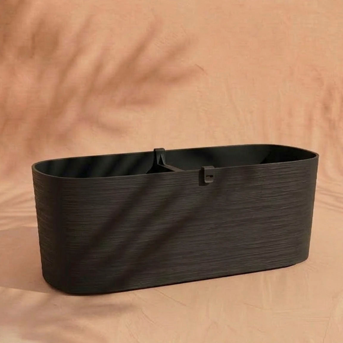 Der Pflanzkasten (Paros | 60 cm) ist ein schwarzes, rechteckiges Pflanzgefäß mit einer subtilen Struktur, ideal für Balkonpflanzen. Entworfen für eine effektive Drainage, ruht er auf einer beigen Oberfläche mit weichen Blattschatten im Hintergrund.