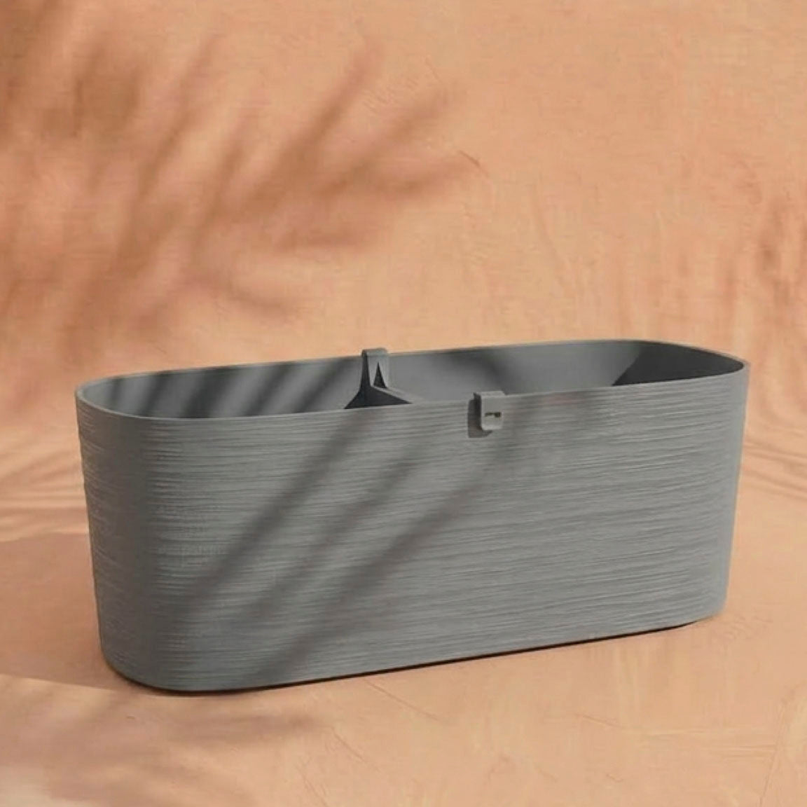 Der graue Pflanzkasten (Paros | 60 cm) mit Mitteltrennwand ruht auf einer pfirsichfarbenen Fläche, die von weichen Blattschatten durchzogen ist - ideal für Balkonpflanzen mit ihrem praktischen Drainagesystem.