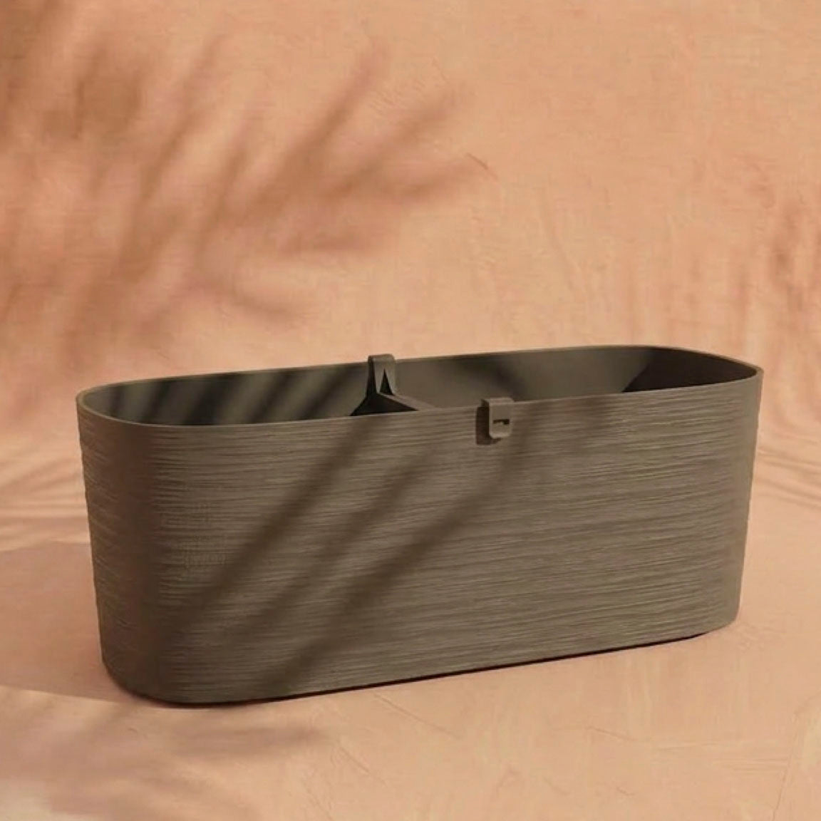 Der Pflanzkasten (Paros | 60 cm) ist ein rechteckiger, olivgrüner Kunststoff-Pflanzkasten mit einem Mittelsteg, ideal für Balkonpflanzen, abgebildet auf einem beigen Hintergrund mit weichen Pflanzenschatten.