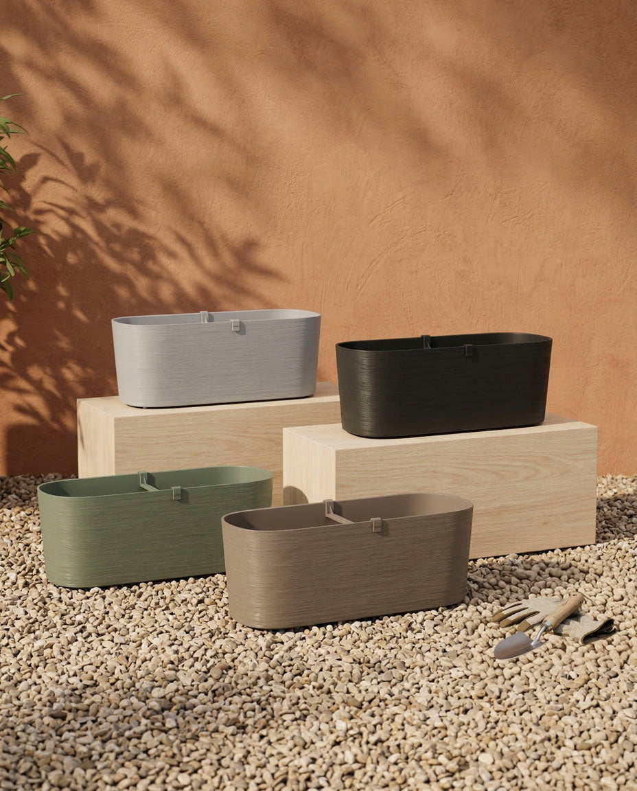 Vier rechteckige Pflanzkästen (Paros | 60 cm) in Grau, Schwarz, Grün und Taupe stehen auf Holzklötzen und Kies zur Entwässerung. In der Nähe stehen Gartengeräte, dahinter wirft eine warme Stuckwand blattreiche Schatten.