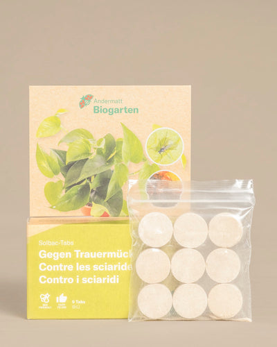 Eine Packung mit neun weissen, runden Solbac-Tabs in einem durchsichtigen Beutel steht vor einer Schachtel mit der Aufschrift "Mittel gegen kleine schwarze Fliegen" zur biologischen Schädlingsbekämpfung, mit Pflanzenbildern und dem Logo des Biogartens Andermatt.