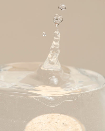 Nahaufnahme einer klaren Flüssigkeit, die aus einem durchsichtigen Glas spritzt, wobei ein dynamischer Wassertropfen eingefangen wird, der die effektive Wirkung von Mitteln gegen kleine schwarze Fliegen durch biologische Schädlingsbekämpfung vor einem neutralen Hintergrund symbolisiert.