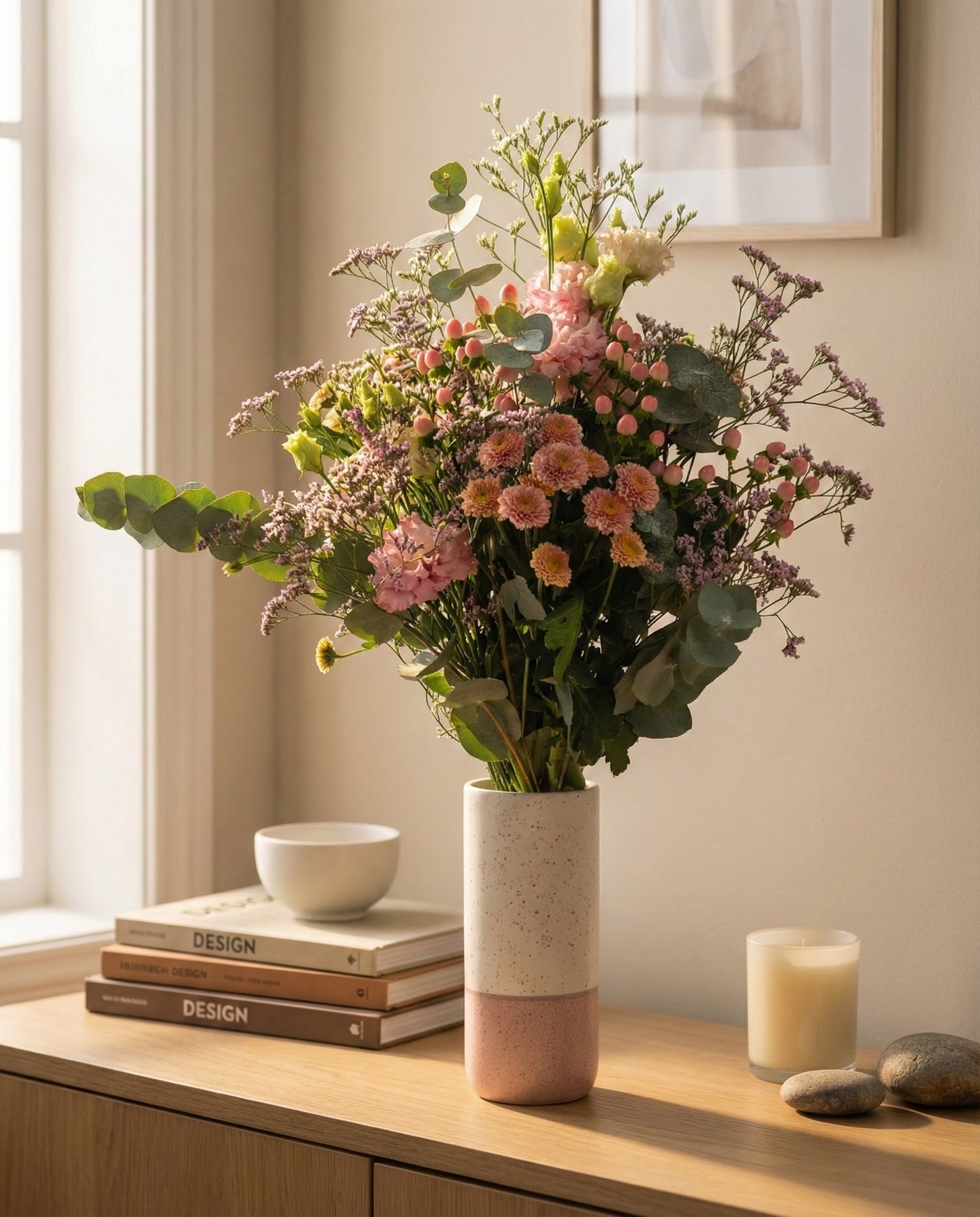 Die Morgenlicht-Vase mit einem handgebundenen Herbstblumenstrauß steht auf einem hölzernen Sideboard zwischen Büchern, einer Schale, Steinen und einer brennenden Kerze und bringt frische Energie in den sanft beleuchteten, gemütlichen Raum.
