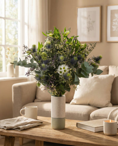 Der Blumenstrauß "Nebelmeer" in Grün und Weiß steht in einer hohen Vase auf dem Couchtisch mit Buch, Tasse, Serviette und brennender Kerze - ein gemütliches Bild im morgendlichen Sonnenschein.