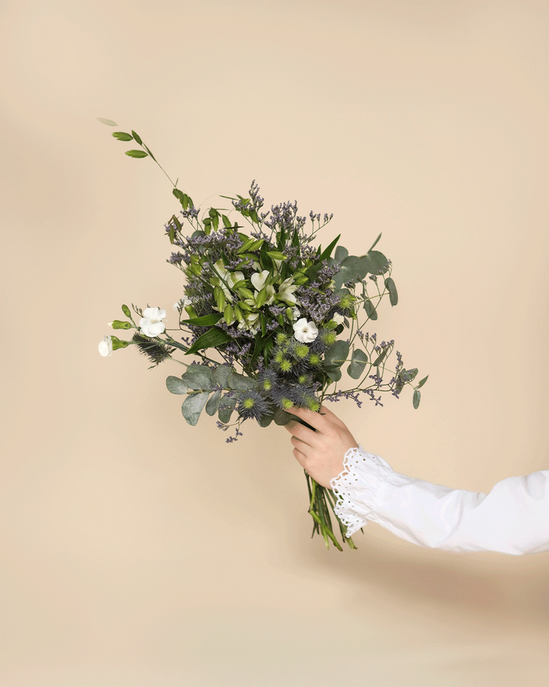 Eine Hand in einer weißen langärmeligen Bluse mit Spitzenmanschetten hält den Strauß "Nebelmeer" - zarte Blüten, grüne Folie und violette Akzente - vor einem einfarbig beigen Hintergrund.