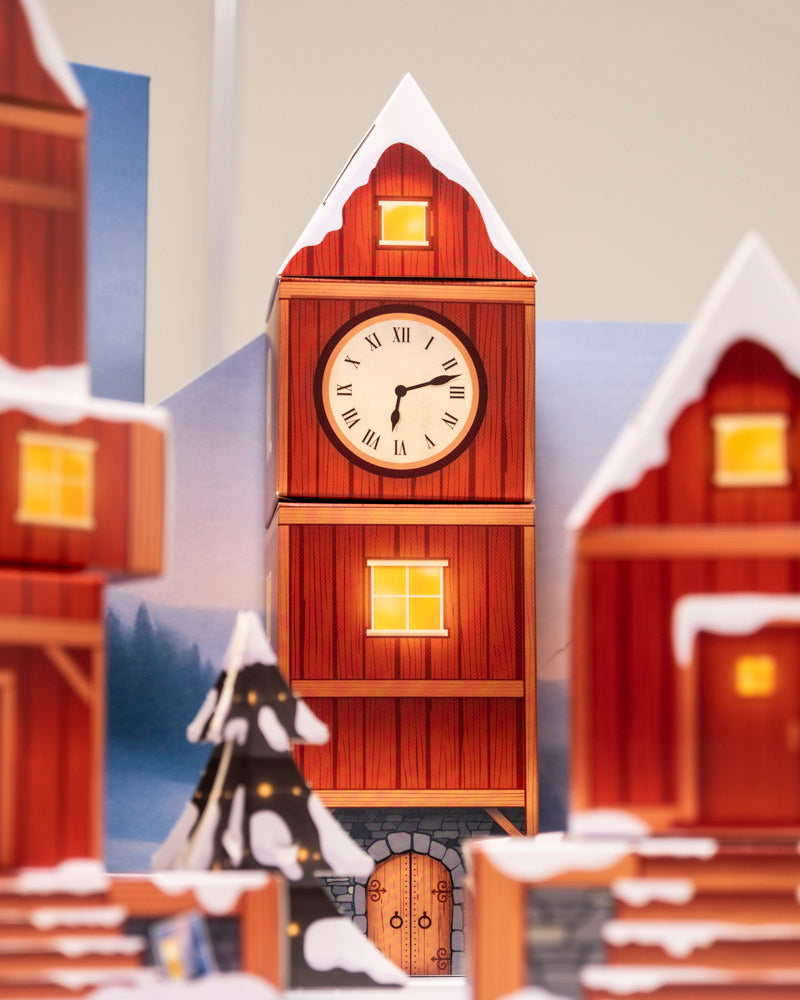 Ein hoher, roter Uhrenturm aus Holz mit Schnee auf dem Dach steht in einer winterlichen Dorfszene, umgeben von festlichen Lichtern und verschneiten Gebäuden - genau wie der Pflanzen-Adventskalender, der für Weihnachtsstimmung sorgt.
