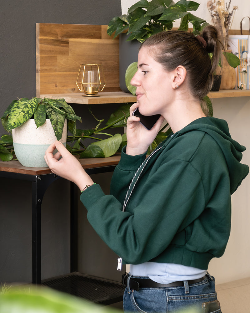 Eine Frau in einem grünen Kapuzenpullover telefoniert, möglicherweise über Pflanzenberatung mit feey, während sie sich um eine Topfpflanze kümmert. Sie berührt sanft eines der Blätter der Pflanze, im Hintergrund sind ein Holzregal mit Dekoartikeln und weiteren Pflanzen zu sehen.