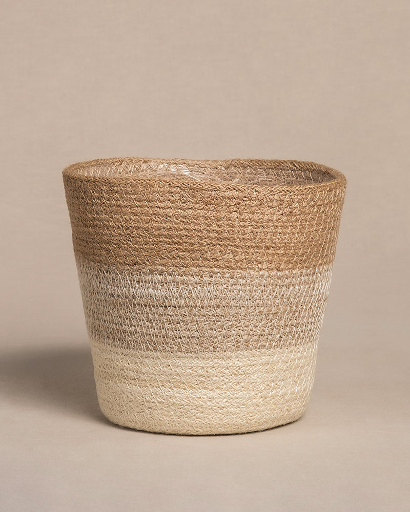 Der Pflanzenkorb aus Jute (Lapis | 18 cm Ø) ist ein geflochtener Korb mit breiter, abgerundeter Oberseite und schmaler Unterseite, der horizontale Farbblöcke in Braun, Beige und Hellcreme aufweist. Er harmoniert wunderbar mit dem neutralen beigen Hintergrund und ergänzt dessen natürliche Töne.