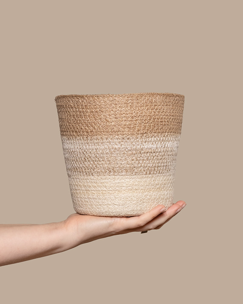 Eine Hand hält einen geflochtenen Korb namens Pflanzenkorb aus Jute (Lapis | 18 cm Ø) mit einem strukturierten Design aus horizontalen Streifen in Creme, Beige und Hellbraun. Er hebt sich vom soliden, neutralen beigen Hintergrund ab und ist perfekt, um Ihrer Einrichtung eine handwerkliche Note zu verleihen.