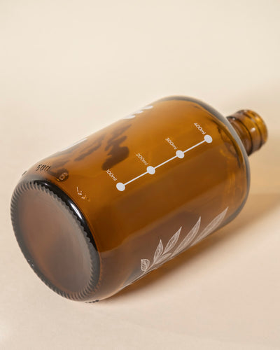 Ein brauner Pflanzensprüher liegt auf der Seite und zeigt eine weiße 100-ml-Messskala und eine botanische Illustration in der Nähe der Basis, alles vor einem weichen beigen Hintergrund.