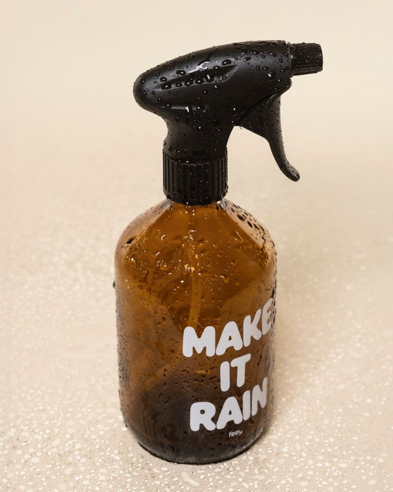 Ein brauner Pflanzensprüher aus Glas mit schwarzer Düse und weißen "MAKE IT RAIN"-Buchstaben steht mit Wassertropfen bedeckt auf einer hellbeigen Oberfläche.