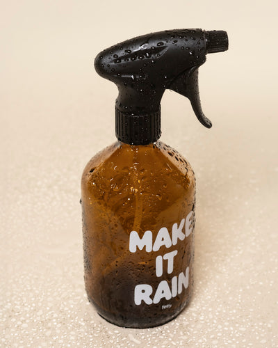 Ein brauner Pflanzensprüher aus Glas mit schwarzer Düse und weißen "MAKE IT RAIN"-Buchstaben steht mit Wassertropfen bedeckt auf einer hellbeigen Oberfläche.