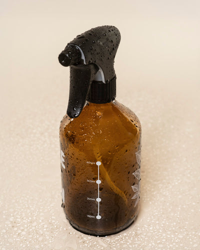 Der Pflanzensprüher aus braunem Glas mit schwarzem Sprühkopf steht aufrechter Position auf einer nassen, hellen Oberfläche. Weiße Messmarkierungen sind deutlich am Produkt sichtbar.