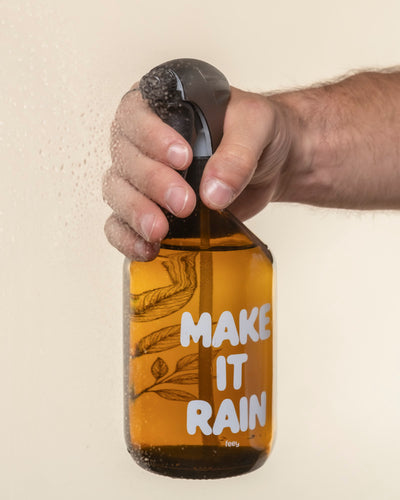Eine Hand sprüht Wasser aus einem bernsteinfarbenen Pflanzensprüher, auf dem in fetten weißen Buchstaben "MAKE IT RAIN" steht, und hält ihn gegen eine beschlagene Glasfläche.