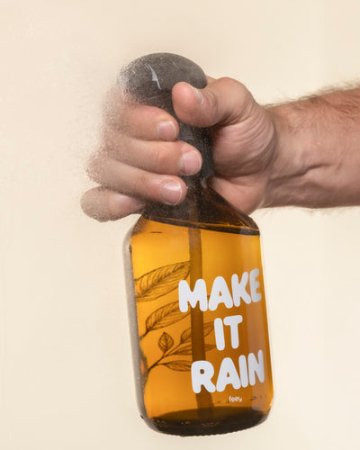 Eine Hand versprüht einen Pflanzensprüher mit der Aufschrift "MAKE IT RAIN" in fetten weißen Buchstaben und zeigt botanische Illustrationen durch das Glas.