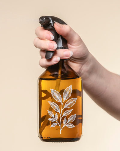 Ein Pflanzensprüher aus braunem Glas mit schwarzer Düse und weißer Pflanzenillustration sprüht Wasser auf Pflanzen. Der sanft beige Hintergrund betont das Design. Ideal zum Besprühen Ihrer Lieblingspflanzen.