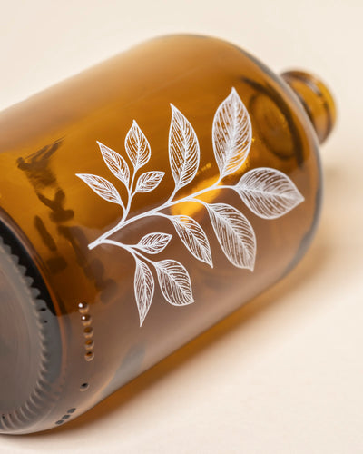 Ein brauner Pflanzensprüher aus Glas mit einer zarten, weißen, blättrigen Zweigdarstellung ruht auf der Seite vor einem hellen, neutralen Hintergrund.
