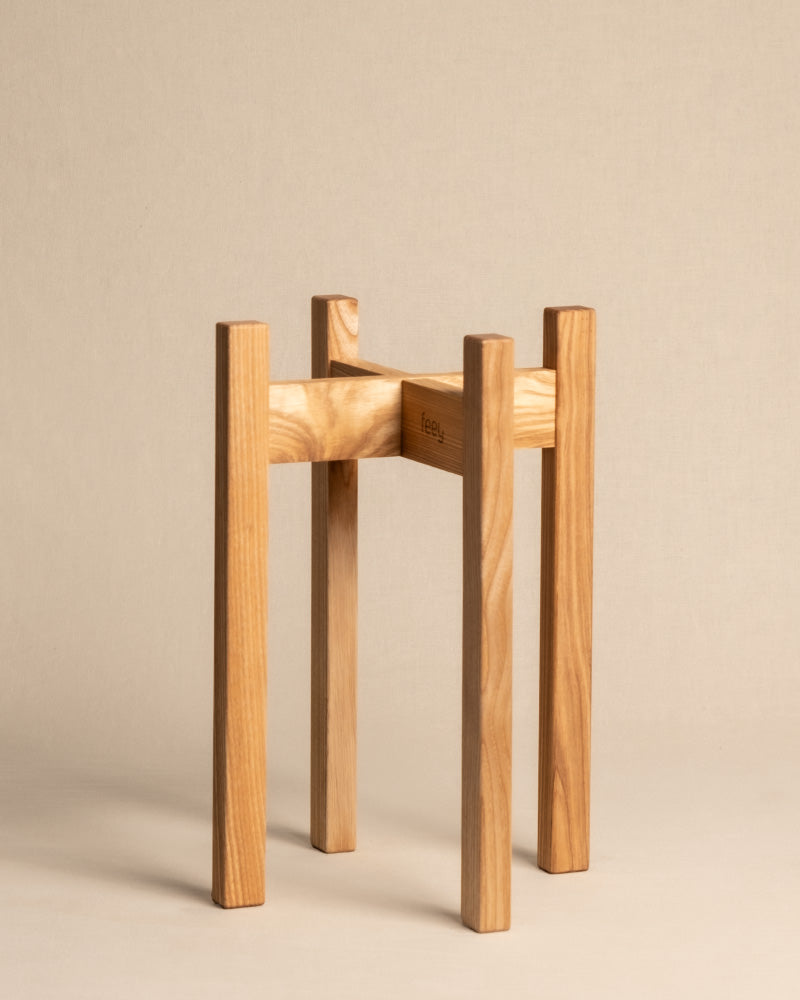 Ein minimalistischer Pflanzenständer (Stelion | 18 cm Ø) mit vier geraden Beinen, gefertigt aus hellem Holz. Das Design ist schlicht und modern und hebt die natürliche Maserung des Holzes vor einem schlichten beigen Hintergrund hervor.