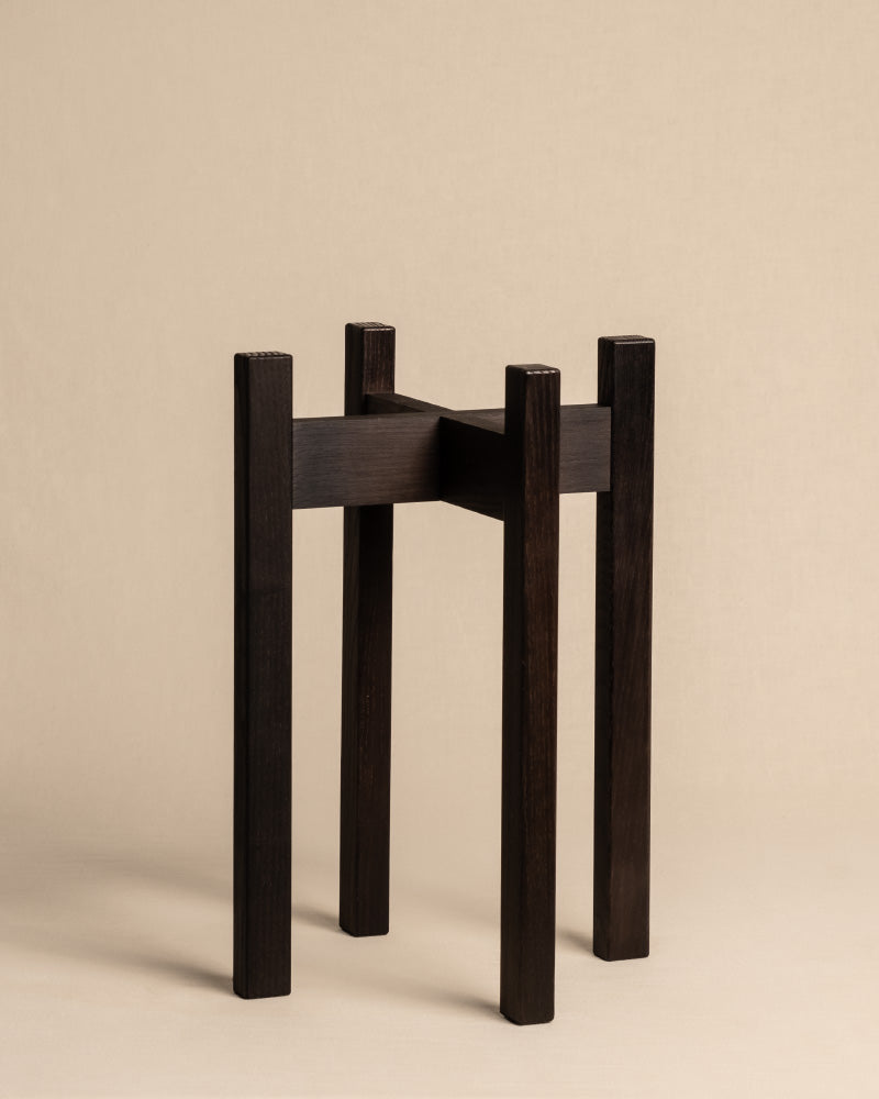 Ein minimalistischer, dunkler Pflanzenständer aus Eschenholz mit runder Basis steht vor einem schlichten beigen Hintergrund. Das schlanke, moderne Design des Ständers mit klaren Linien macht ihn zu einem perfekten Unikat in jedem Raum.