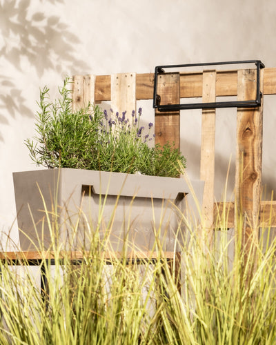 Holzkiste mit einem beigen Pflanzkasten für Geländer (Bruges | 55 cm), gefüllt mit Lavendel und Grünpflanzen, auf einem Hintergrund aus Holzpaletten. Sonnenlicht wirft Schatten von Blättern an die Wand, und hohes Gras im Vordergrund verleiht dem Ganzen eine natürliche, rustikale Note.