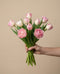 Eine Hand hält einen Strauß Tulpen "Liebesgedicht" - rosafarbene und weiße Tulpen mit grünen Stielen und Blättern - vor einem einfarbig beigen Hintergrund.