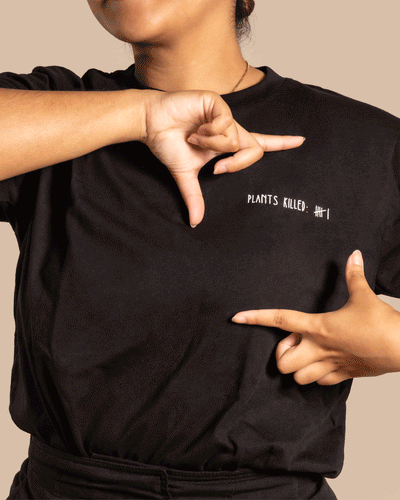 Eine Person trägt ein schwarzes Pflanzenshirt mit dem Text „PLANTS KILLED: IIII“. Die Person macht Handbewegungen, die den Text auf dem Shirt einrahmen, anzeigen oder hervorheben. Perfekt für Pflanzenfans und alle, die Wert auf ökologisch nachhaltige Kleidung legen.