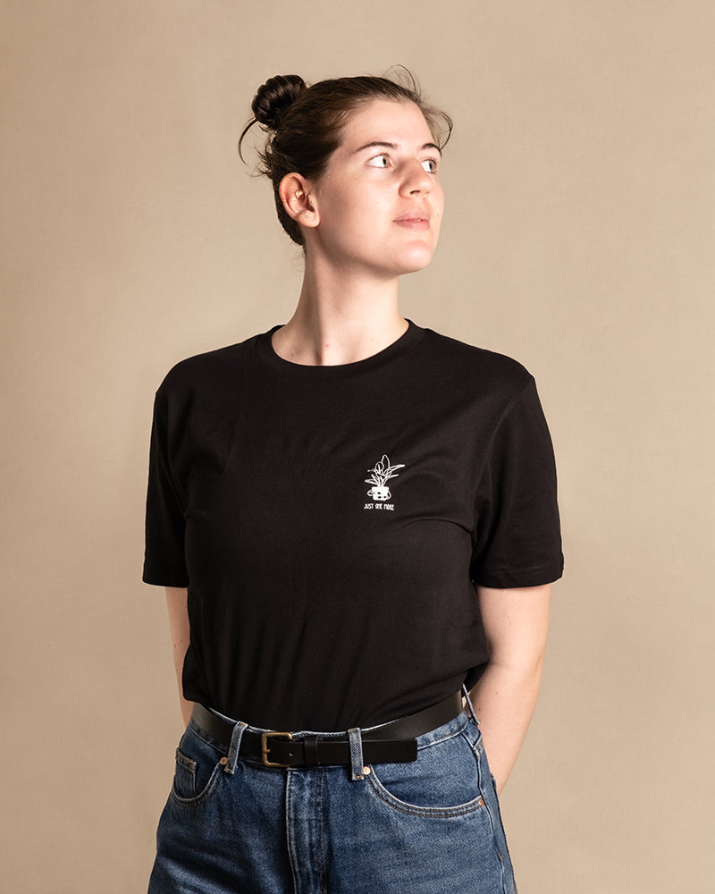 Eine Person mit zurückgekämmten Haaren blickt zur Seite. Sie trägt ein schwarzes Plant Shirt aus BIO-Baumwolle mit kleiner Grafik und Text auf der linken Brustpartie und blaue Jeans. Sie hat einen neutralen Gesichtsausdruck, die Hände sind hinter dem Rücken und der Hintergrund ist schlicht beige.