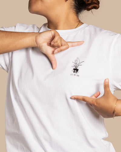 Eine Person trägt ein weißes T-Shirt aus BIO-Baumwolle und zeigt auf das kleine gestickte Motiv auf dem Shirt, das eine Topfpflanze und den Text „Just One Piece“ zeigt. Das Gesicht der Person ist nicht zu sehen. Sie lenkt mit ihren Händen die Aufmerksamkeit auf das Motiv – perfekt für Plant Shirts.