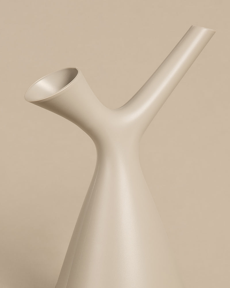 Eine minimalistische, beige Gießkanne in modernem Design mit einer glatten, glatten Oberfläche. Diese Plunge-Gießkanne verfügt über einen einzigartigen Y-förmigen Ausguss und eine längliche, schmale Düse, die oben hervorsteht und sich von einem passenden beigen Hintergrund abhebt.